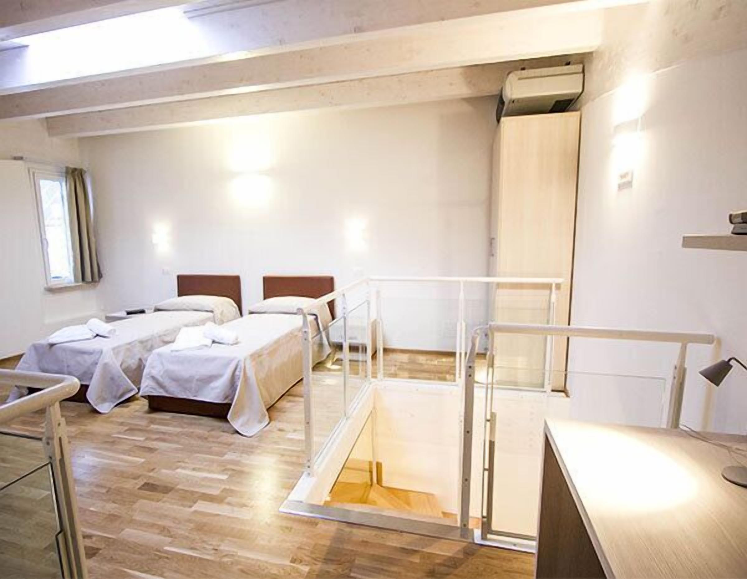 A hotel room with two beds, a wooden floor, a window with curtains, and a white wall.