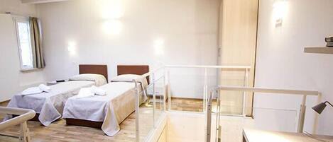 A hotel room with two beds, a wooden floor, a window with curtains, and a white wall.