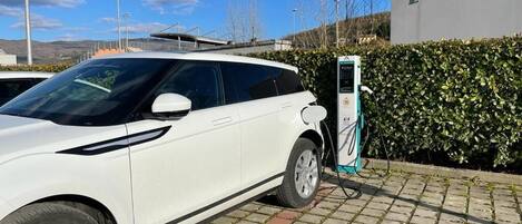 Un vehículo eléctrico blanco cargando en una estación, con un seto y un edificio al fondo.