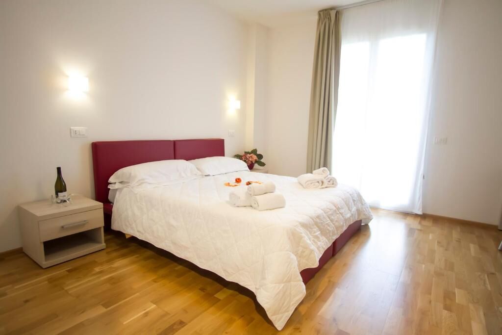 A neatly arranged bedroom with a wooden floor, a bed with white bedding, a nightstand with a bottle, and a window with curtains.