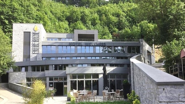 A modern building with a glass facade, outdoor seating area, and a stone retaining wall. - Hotel Wellness Eau de Roche (Durbuy)