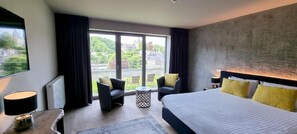 A modern hotel room with a large bed, two armchairs, a small table, and a view of a cityscape through large windows. - Hotel Wellness Eau de Roche (Durbuy)