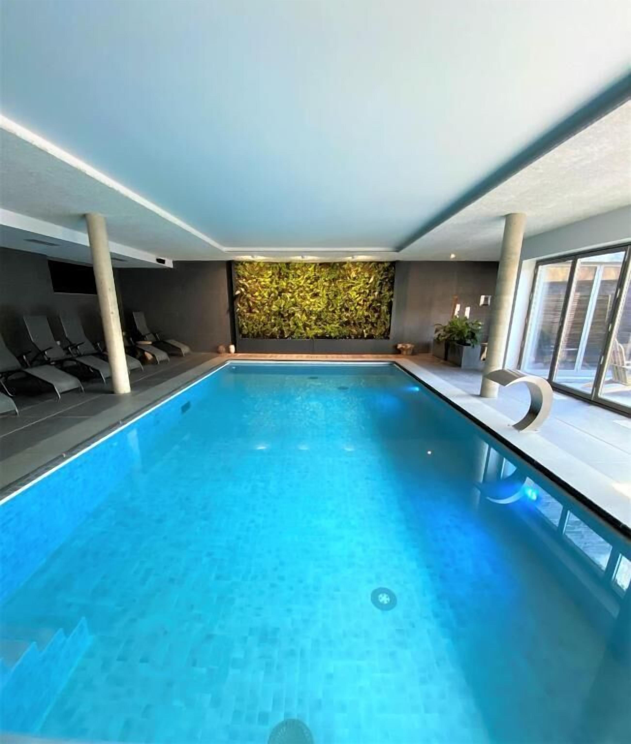 An indoor swimming pool with a clear view of the ceiling and walls, a seating area with chairs, and a decorative wall with green foliage.