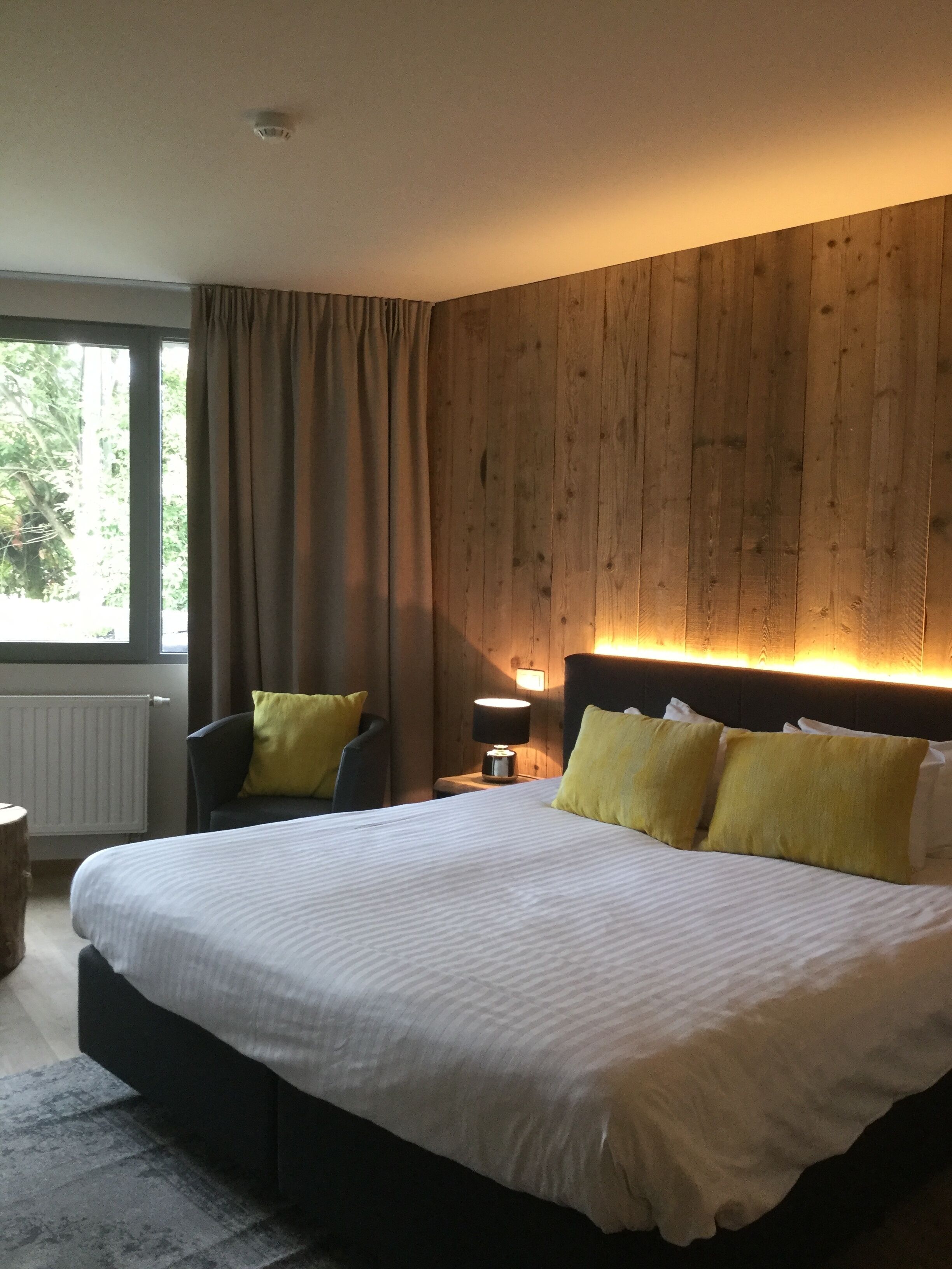 Een slaapkamer met een groot bed, houten paneelwanden, een nachtkastje met een lamp en een raam met gordijnen.