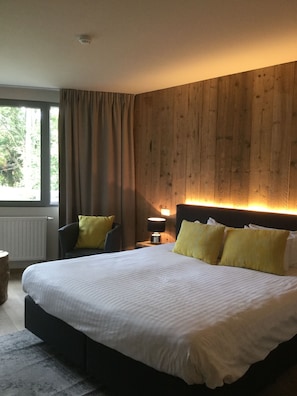 A bedroom with a large bed, wooden panel walls, a nightstand with a lamp, and a window with curtains.