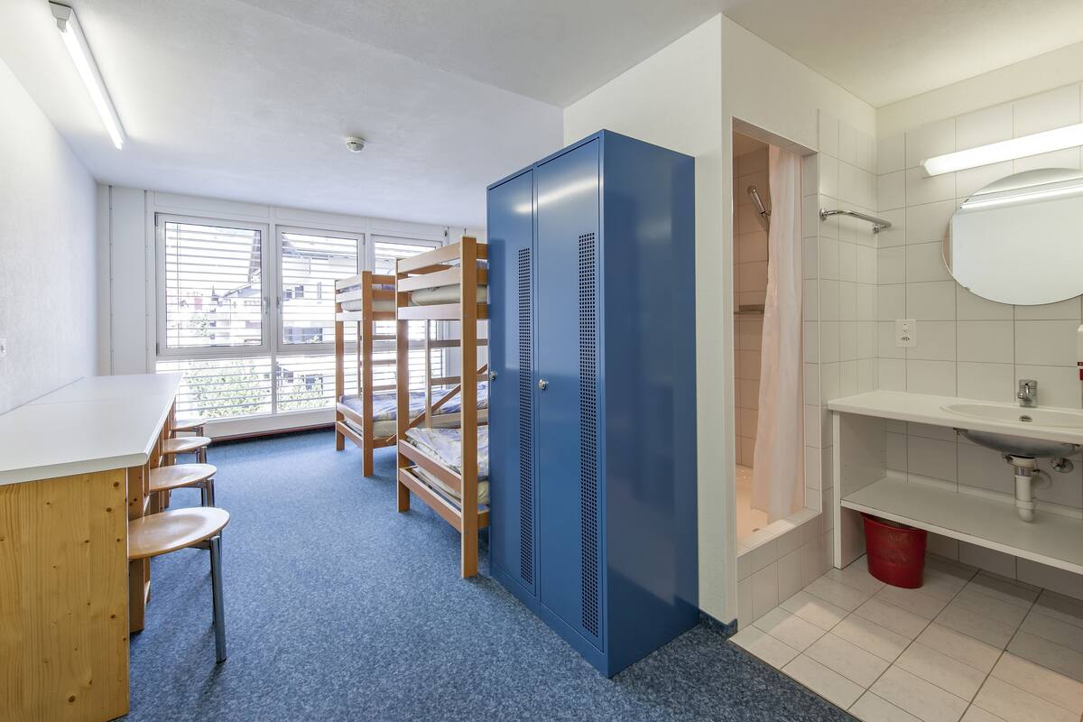 Een kamer met stapelbedden, een bureau, stoelen, een blauwe kast, een badkamer met wastafel en spiegel, en een raam met jaloezieën.