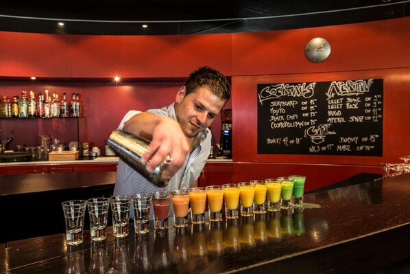 A bartender preparing cocktails at a bar with a menu board in the background. - Hostel INN LODGE (Celerina-Schlarigna)