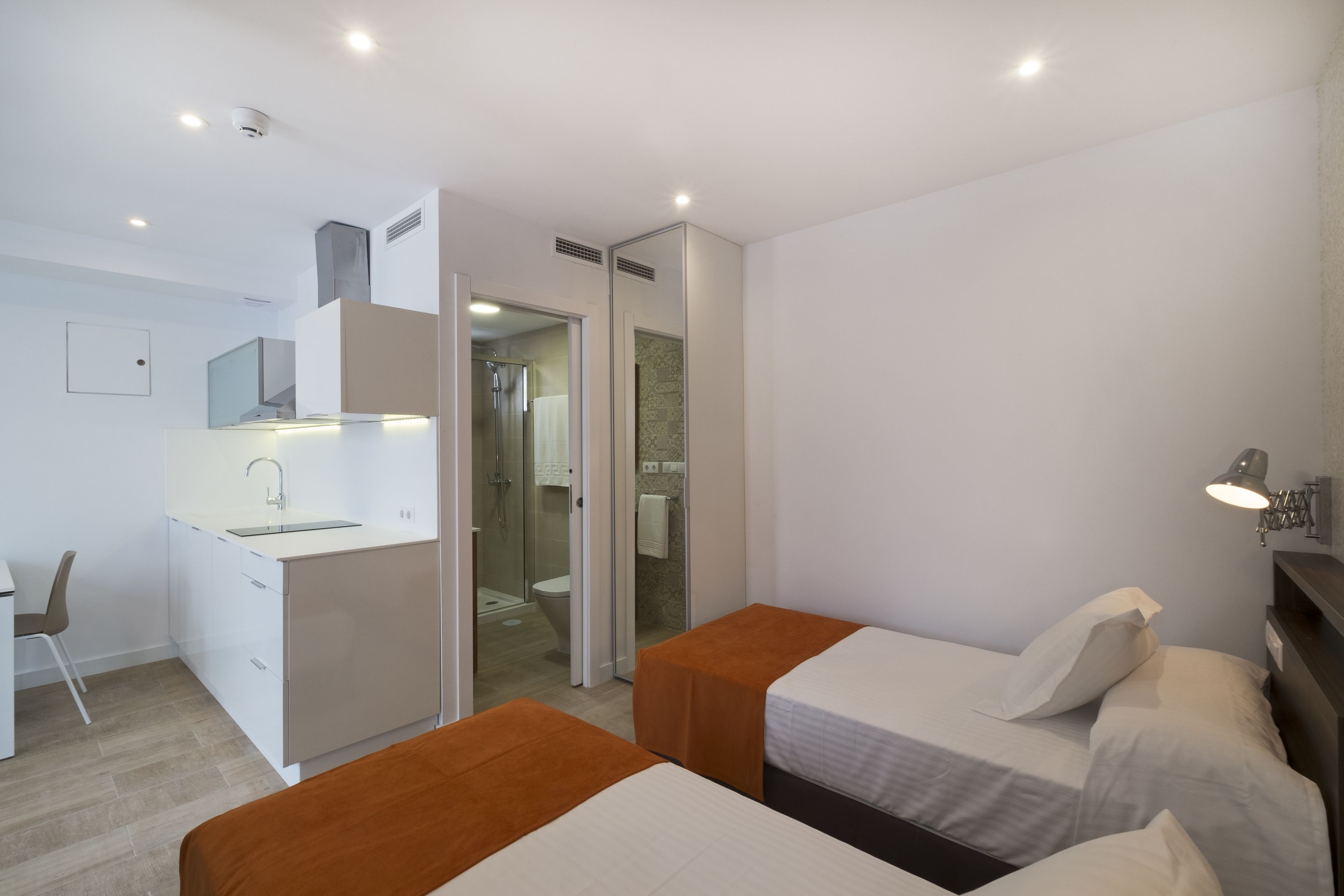 Ein modernes Hotelzimmer mit zwei Einzelbetten, einem Schreibtisch mit Stuhl, einer Duschkabine und einer kleinen Küchenzeile.