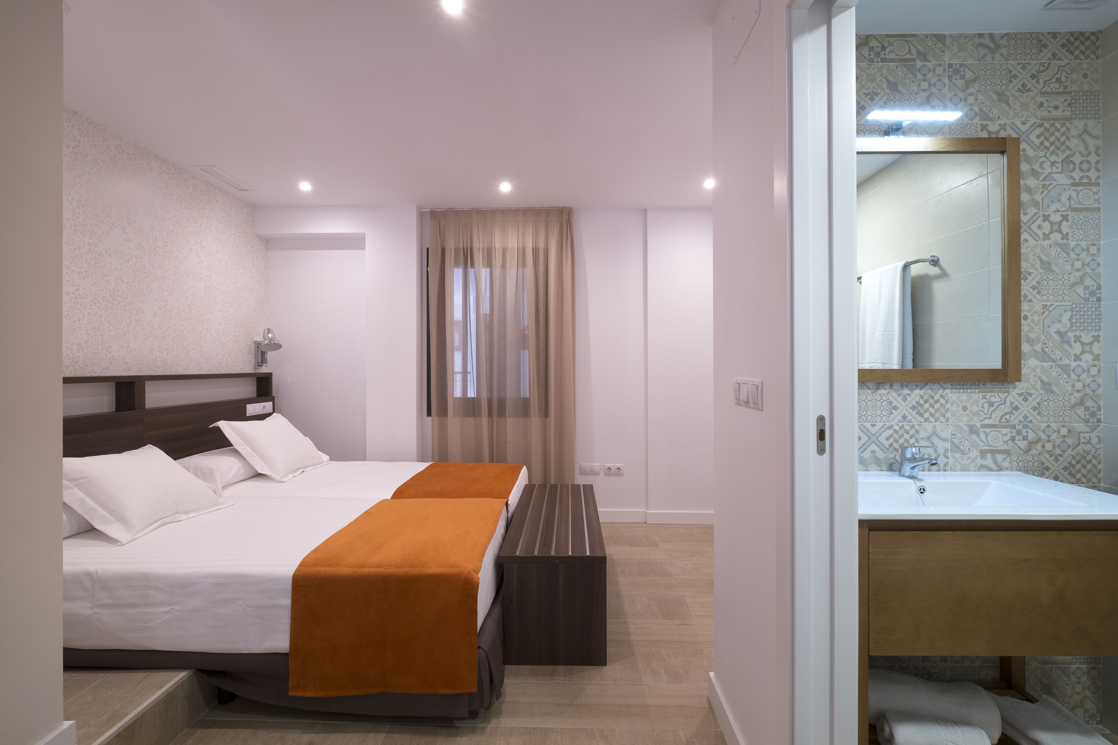 A modern hotel room with a large bed, a bench, a bathroom with a sink and mirror, and patterned walls.