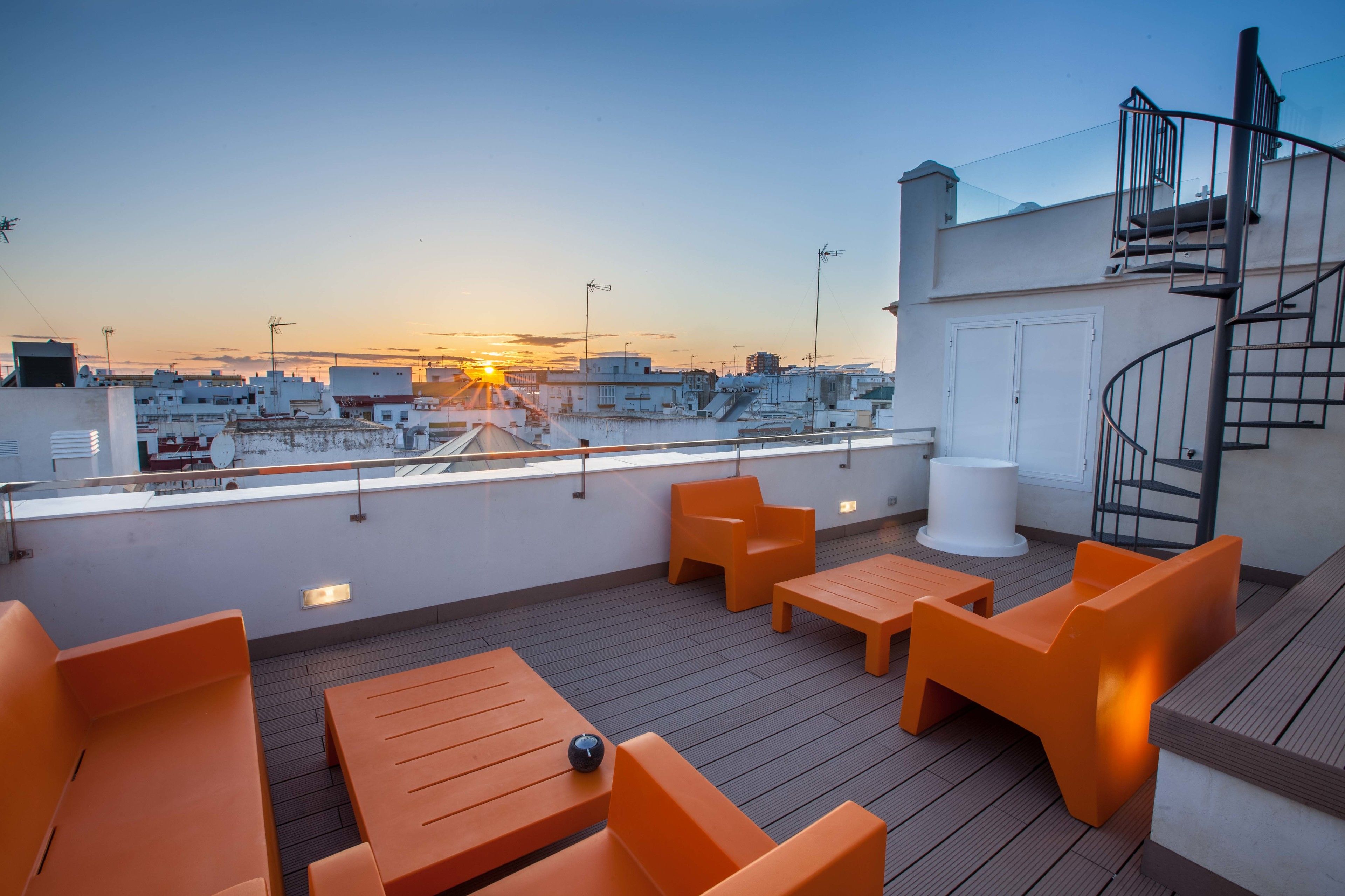 A rooftop terrace with orange furniture, a white cylindrical object, and a view of the cityscape during sunset.