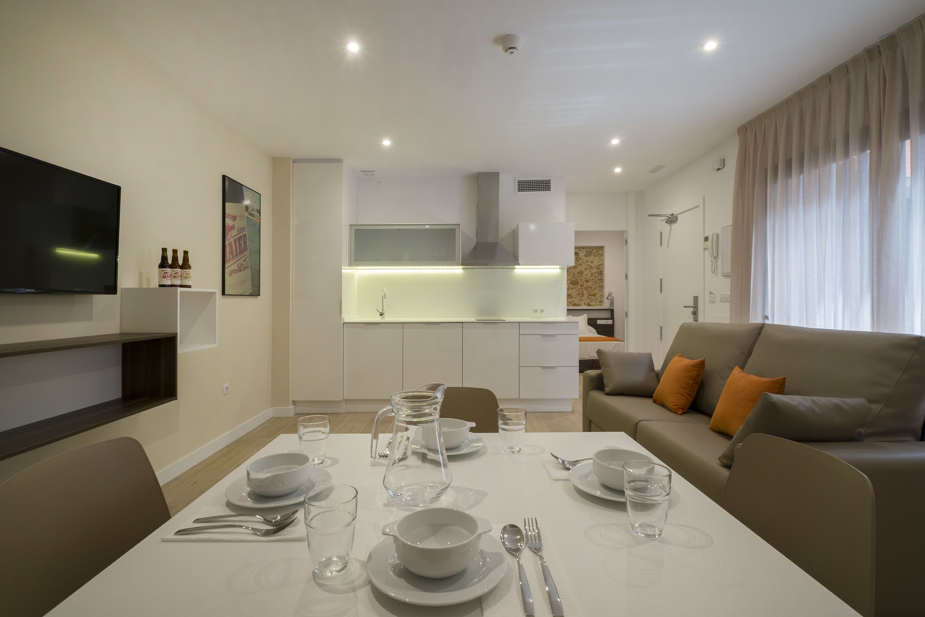 Una sala moderna con una mesa de comedor preparada para cuatro, un sofá con cojines naranjas y una cocina al fondo.