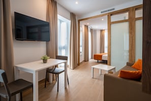 A modern hotel room with a flat-screen TV, a dining table with chairs, a sofa with orange cushions, and a bed in the background. - Apartamentos Maier (Cádiz)