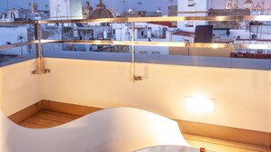 A modern rooftop terrace with a white lounge chair, a small table with bottles, and a cityscape view in the background. - Apartamentos Maier (Cádiz)