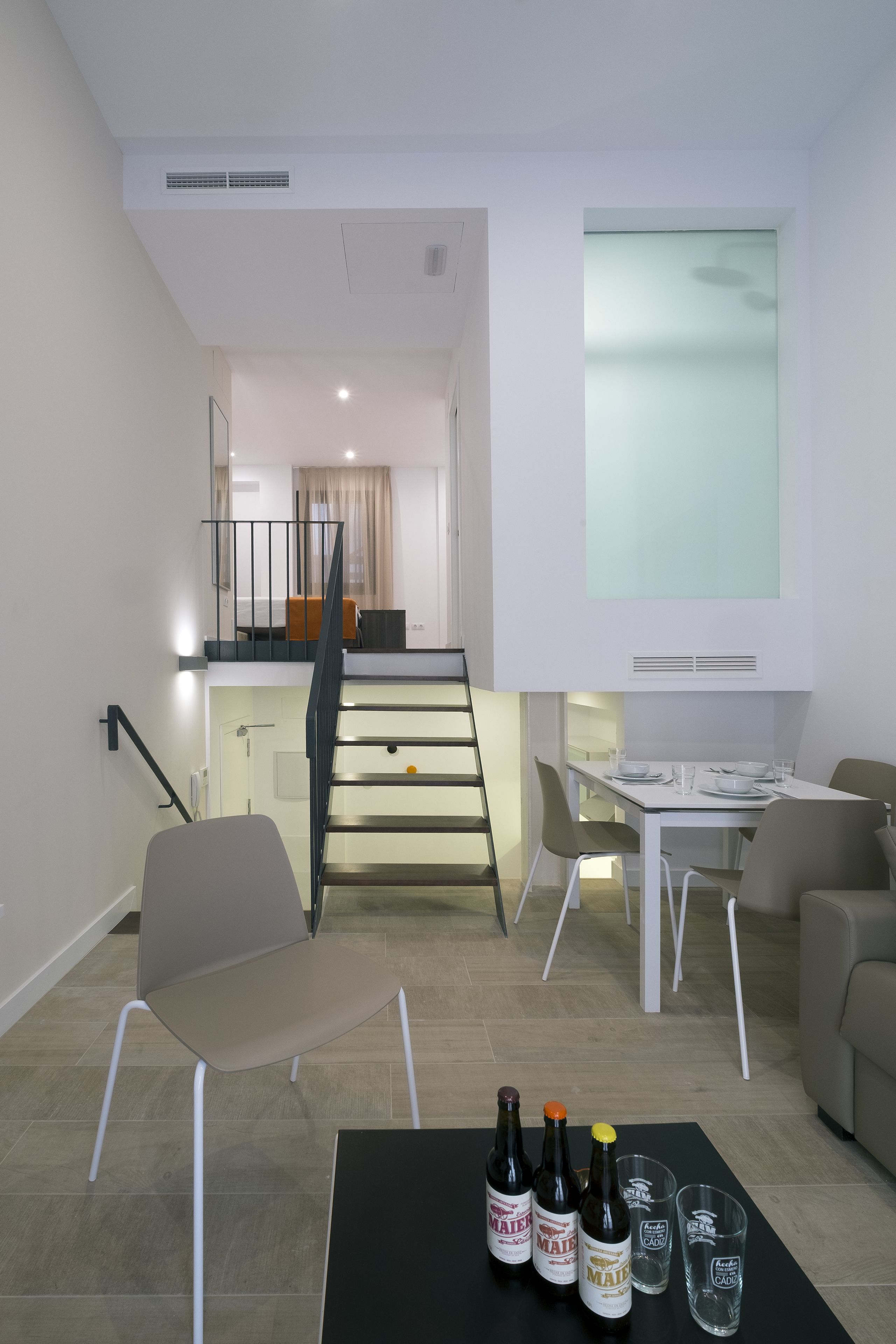 A modern living room with a dining table, chairs, and a staircase leading to an upper floor. There are bottles and glasses on the table.