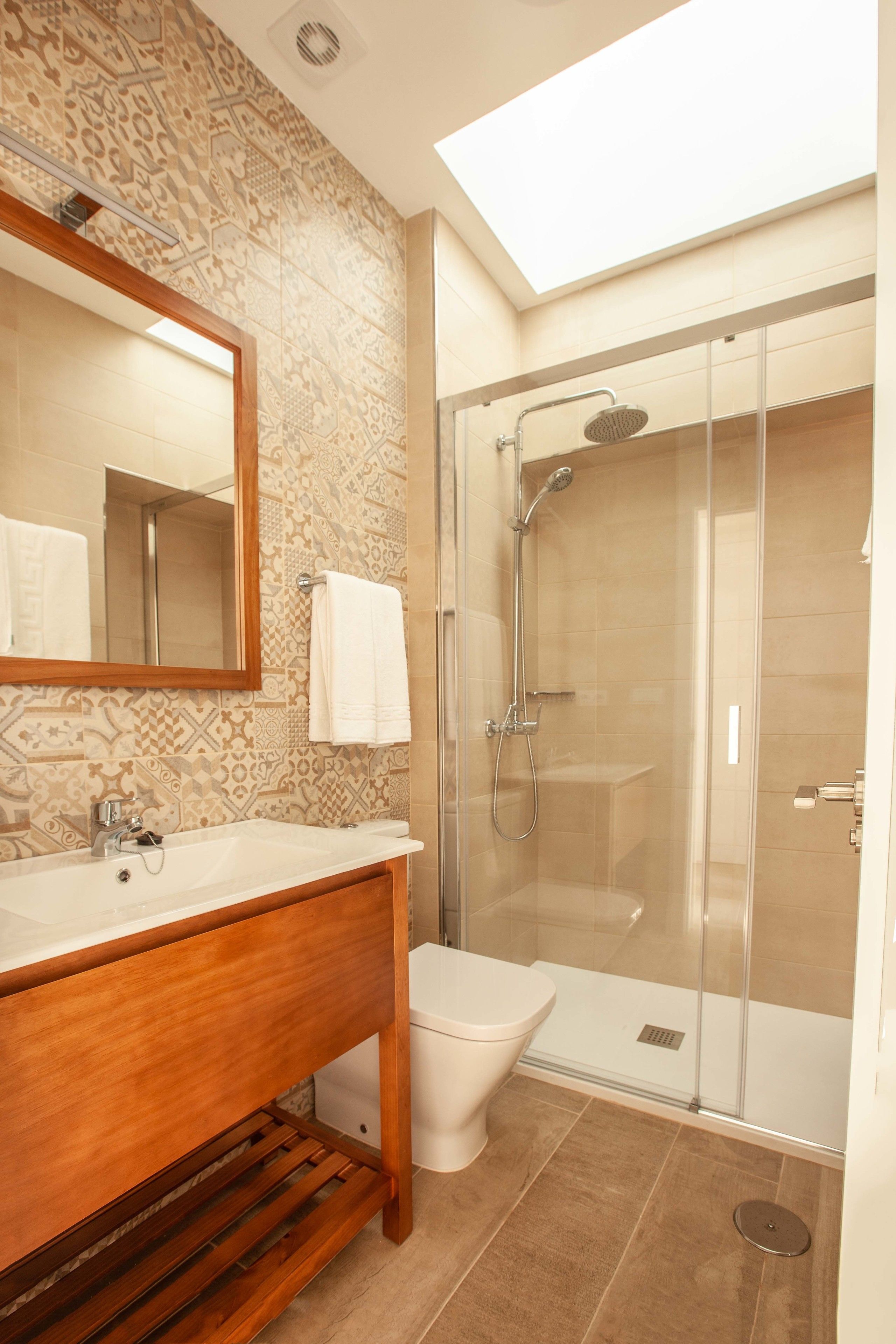 Un baño con tocador de madera, inodoro blanco, ducha con cerramiento de vidrio y tragaluz.