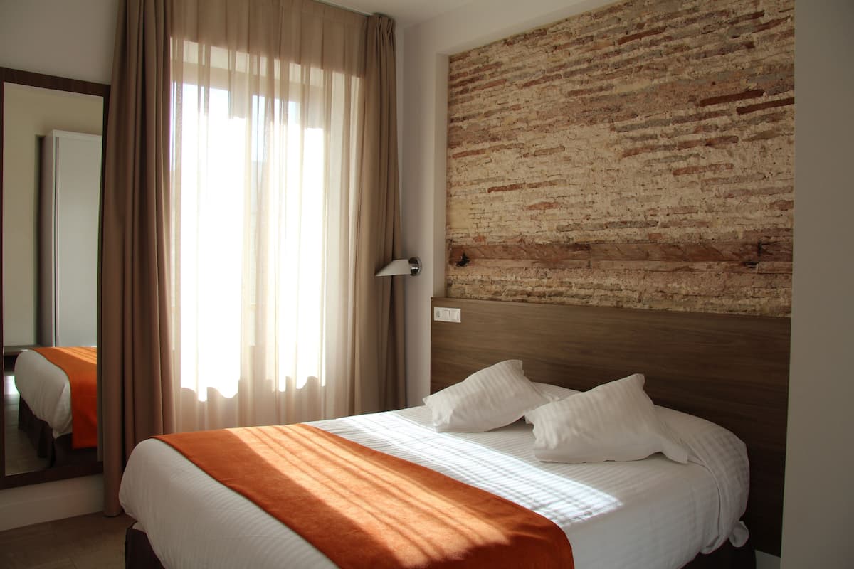 Ein Hotelzimmer mit einem großen Bett, einer Wand mit Backsteinverkleidung und einem Fenster mit durchsichtigen Vorhängen.