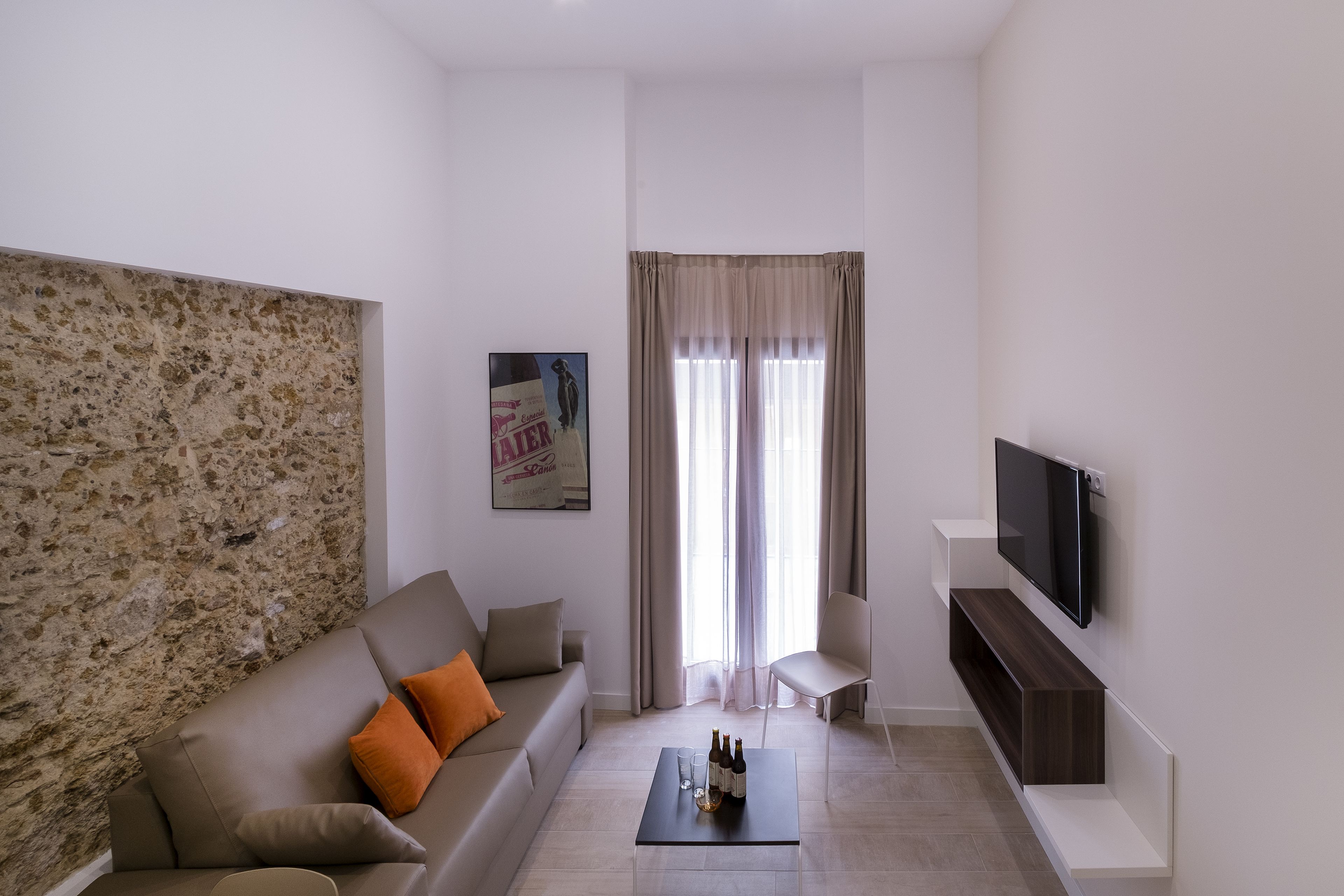 Een moderne woonkamer met een stenen accentmuur, een bank met oranje kussens, een salontafel met flessen, een aan de muur bevestigde televisie en een groot raam met gordijnen.