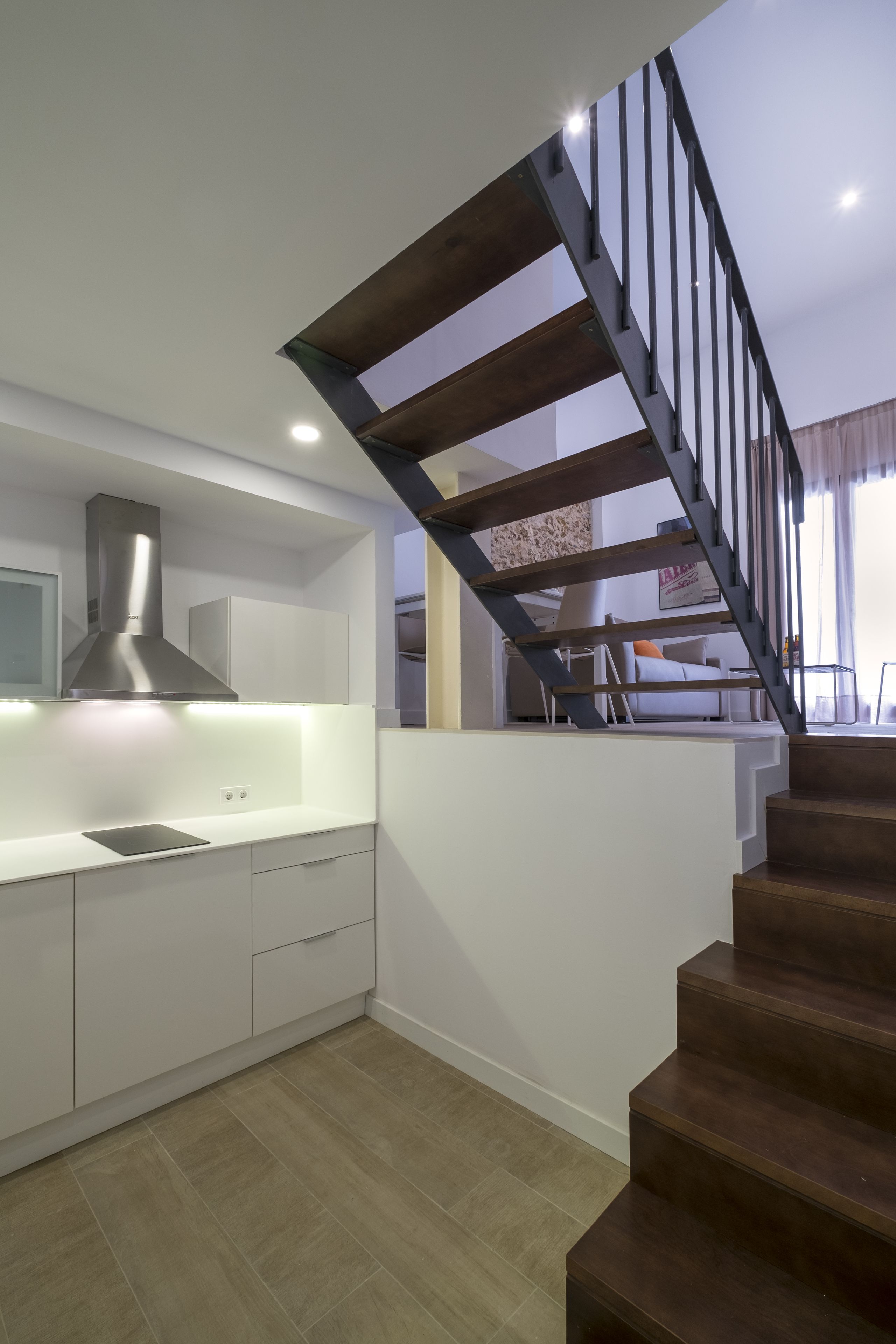 Una cocina moderna con armarios blancos, una campana extractora de acero inoxidable y una escalera de madera con barandillas negras.