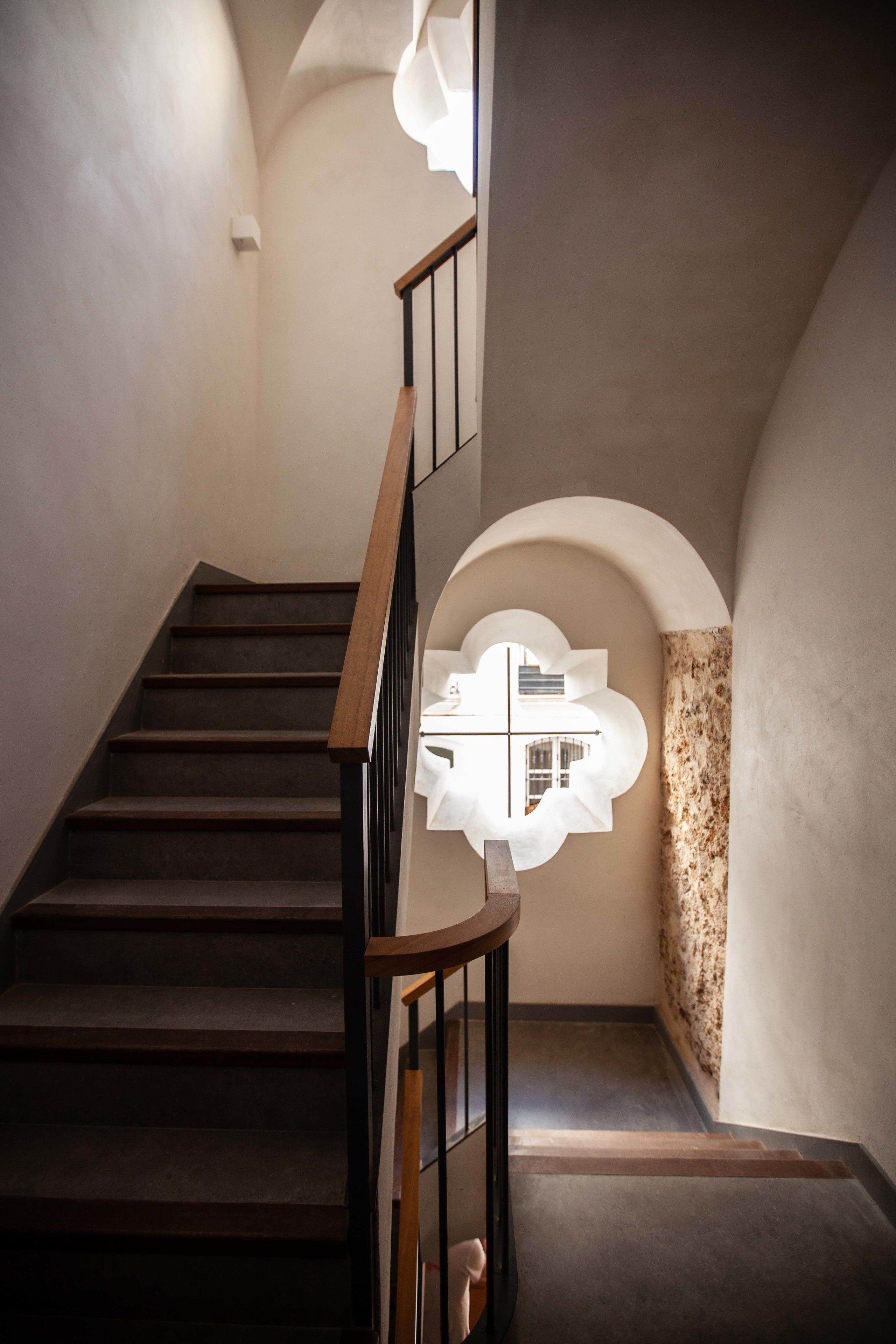 A staircase with wooden steps and a black railing, leading to a landing with a textured wall and a circular window.