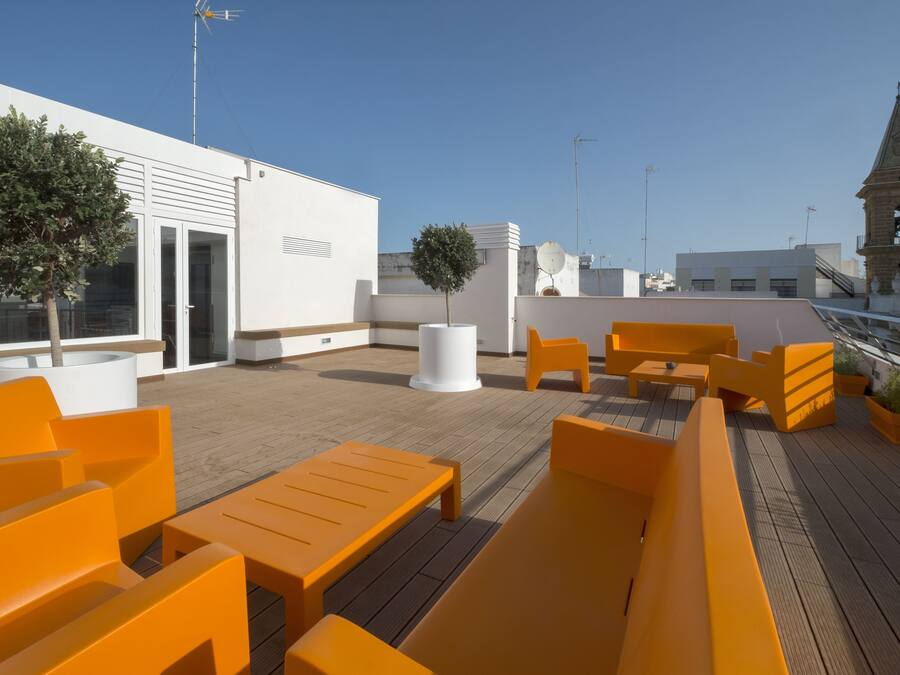 Una terraza en la azotea con asientos anaranjados, un edificio blanco y una torre de reloj al fondo.