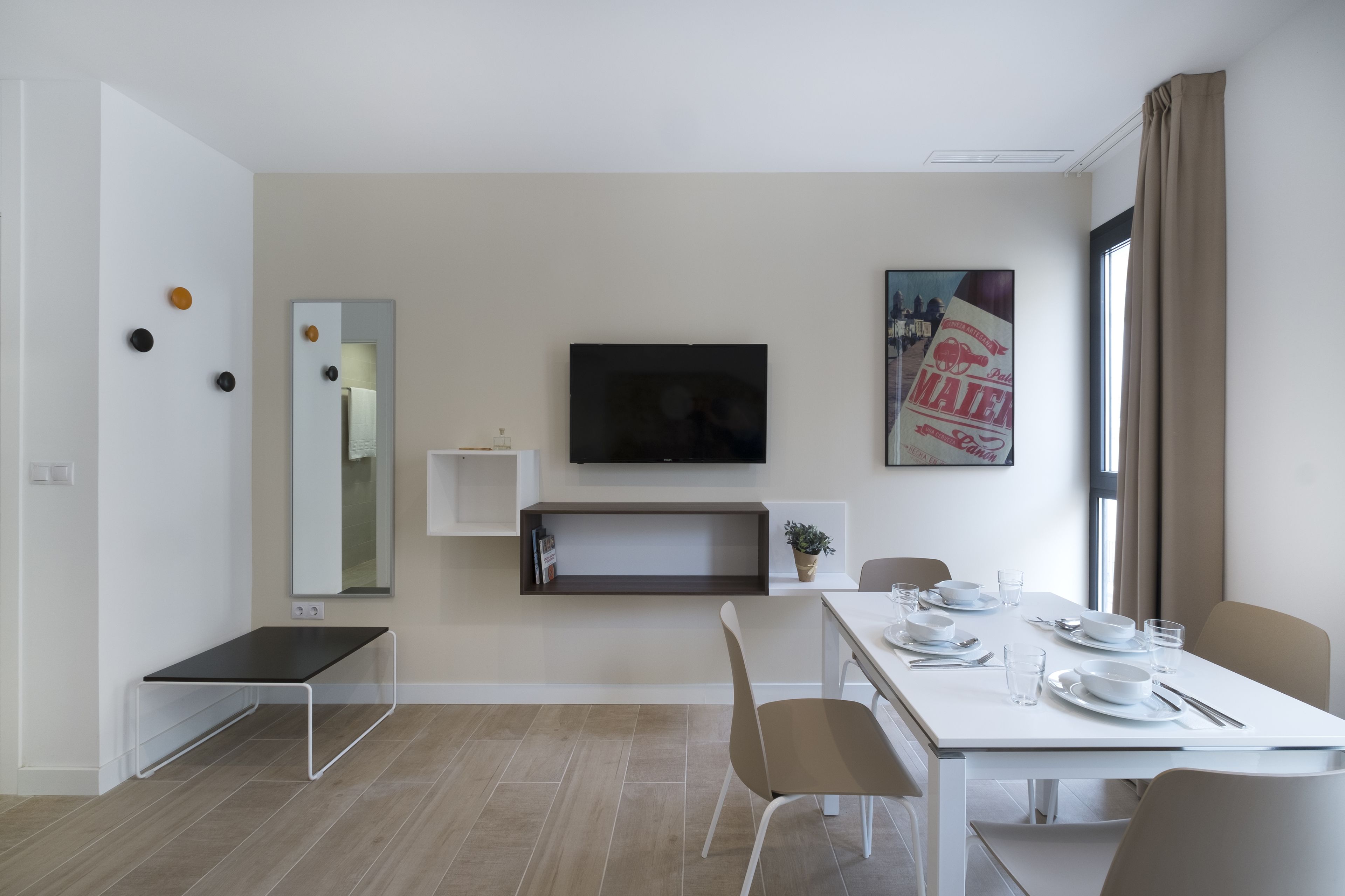 Een moderne, goed verlichte kamer met een eettafel gedekt voor twee personen, een flatscreen televisie aan de wand en een groot raam met gordijnen.