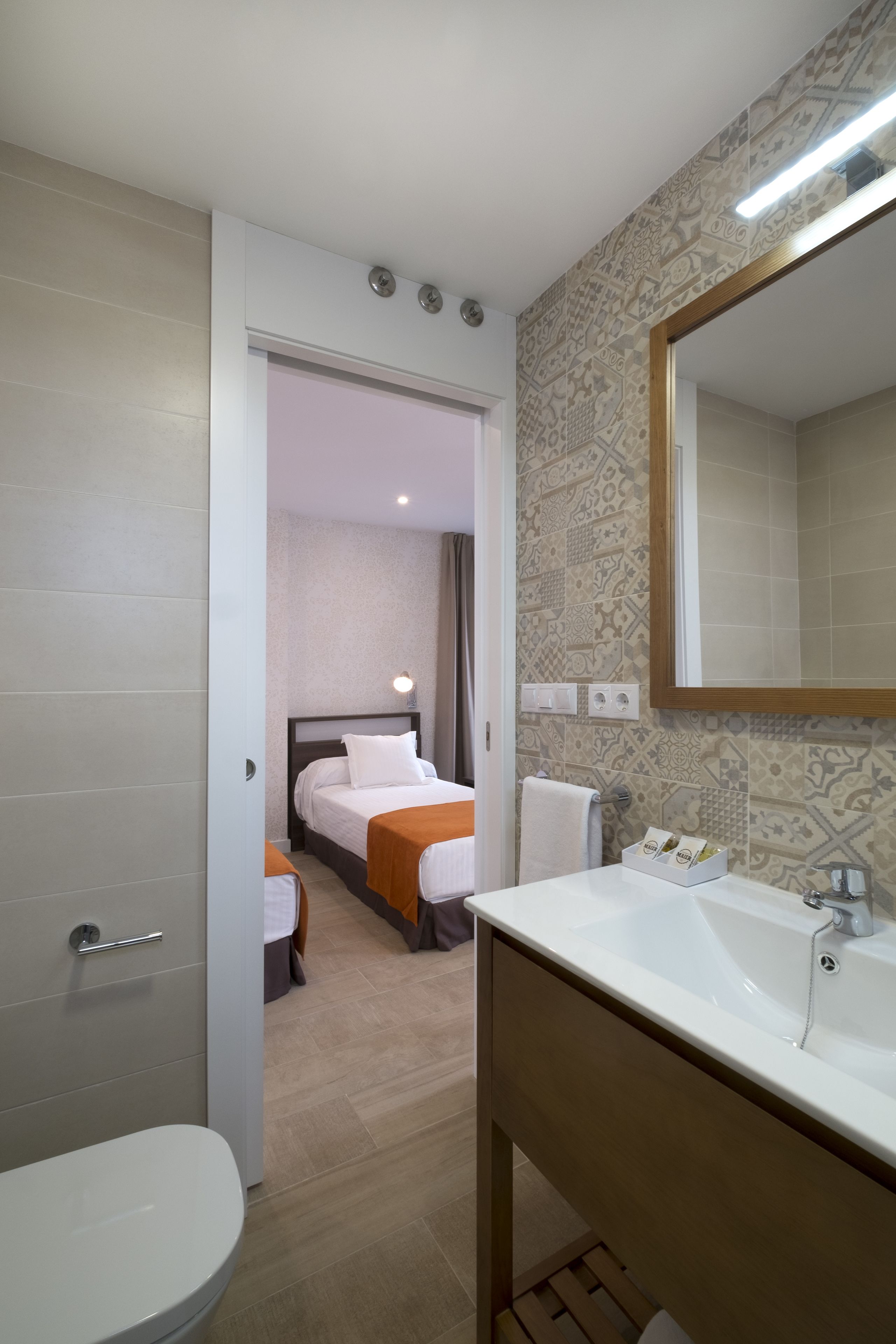Ein Hotelbad mit Waschbecken, Spiegel und Toilette. Im angrenzenden Raum ist ein Bett sichtbar.