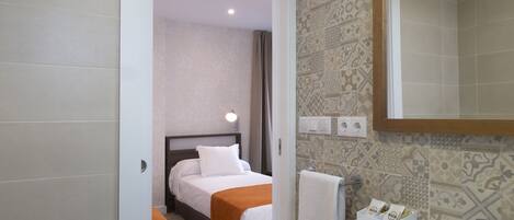 A hotel bathroom with a white sink, a mirror, and a toilet. There is a bed visible in the adjacent room.