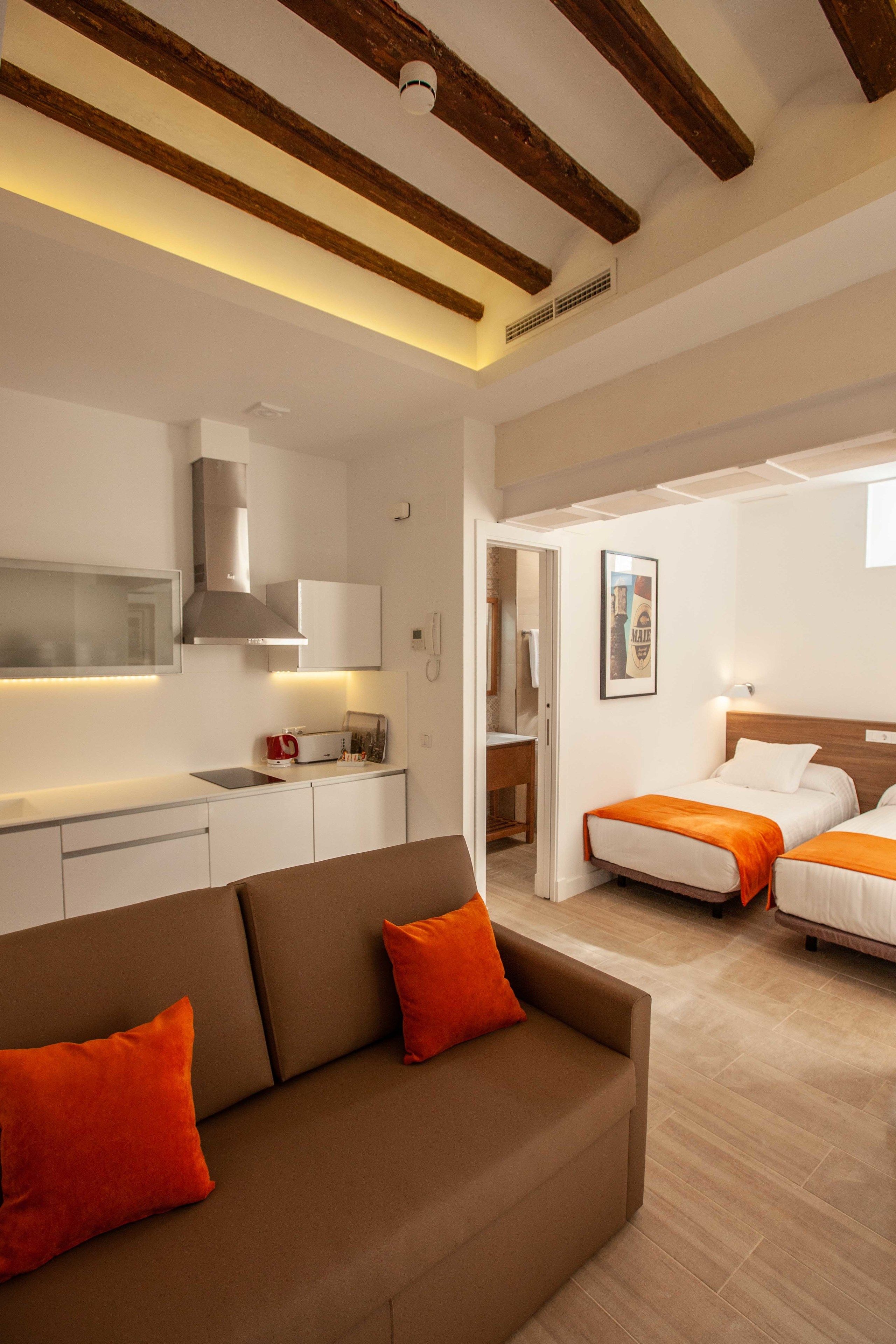 A modern hotel room with a kitchenette, two beds, a sofa, and a bathroom visible through an open door.