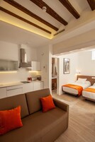 A modern hotel room with a kitchenette, two beds, a sofa, and a bathroom visible through an open door.