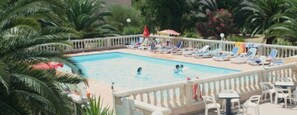 A swimming pool with lounge chairs and umbrellas, surrounded by palm trees and a white fence. - Résidence Thalassa (Calvi)