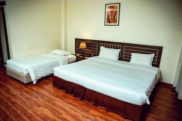 A hotel room with two beds, wooden flooring, a nightstand with a lamp, and a painting on the wall.