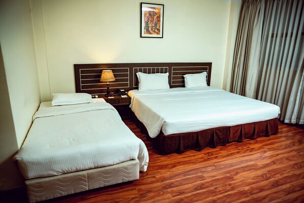 A hotel room with two single beds, a wooden floor, a nightstand with a lamp, and a painting on the wall.