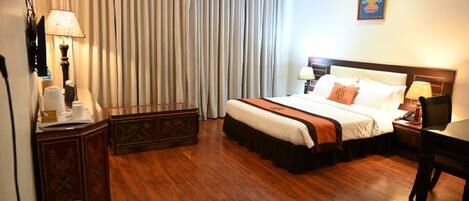 A hotel room with a large bed, a desk, a chair, a lamp, a trash bin, and a curtain.