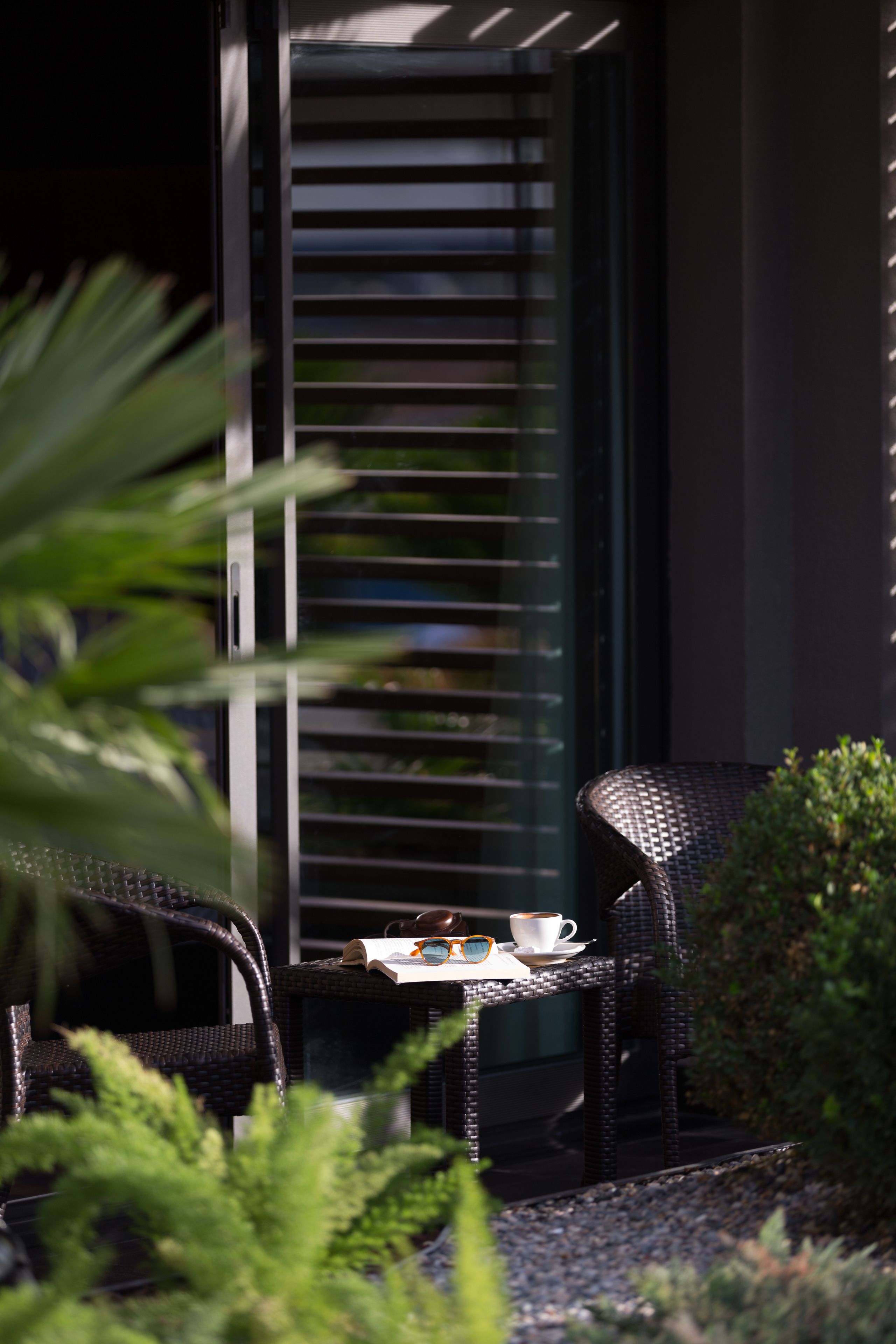 Una silla de mimbre y una mesa con una taza y gafas de sol, frente a un edificio con ventana y persianas.