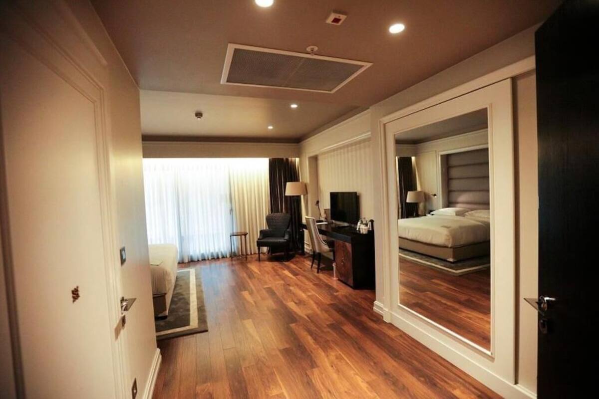 Une chambre d’hôtel avec un plancher en bois, un lit, un bureau avec une chaise, une télévision et une fenêtre avec des rideaux.