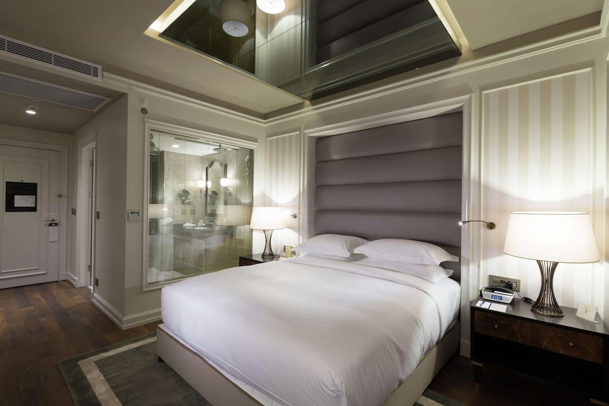 Une chambre d’hôtel avec un grand lit, des lampes de chevet, une table de chevet, un miroir et une fenêtre avec des stores.