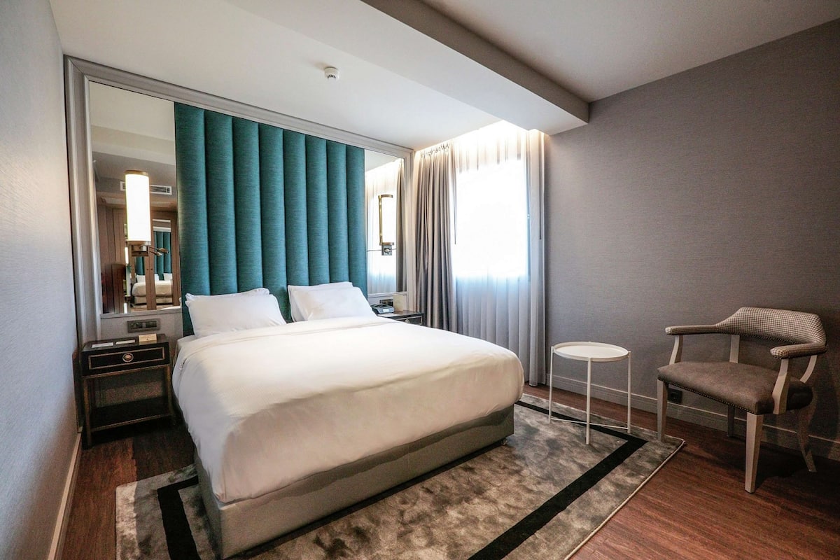 Une chambre d’hôtel moderne avec un grand lit, une petite table ronde, une chaise et un grand miroir.