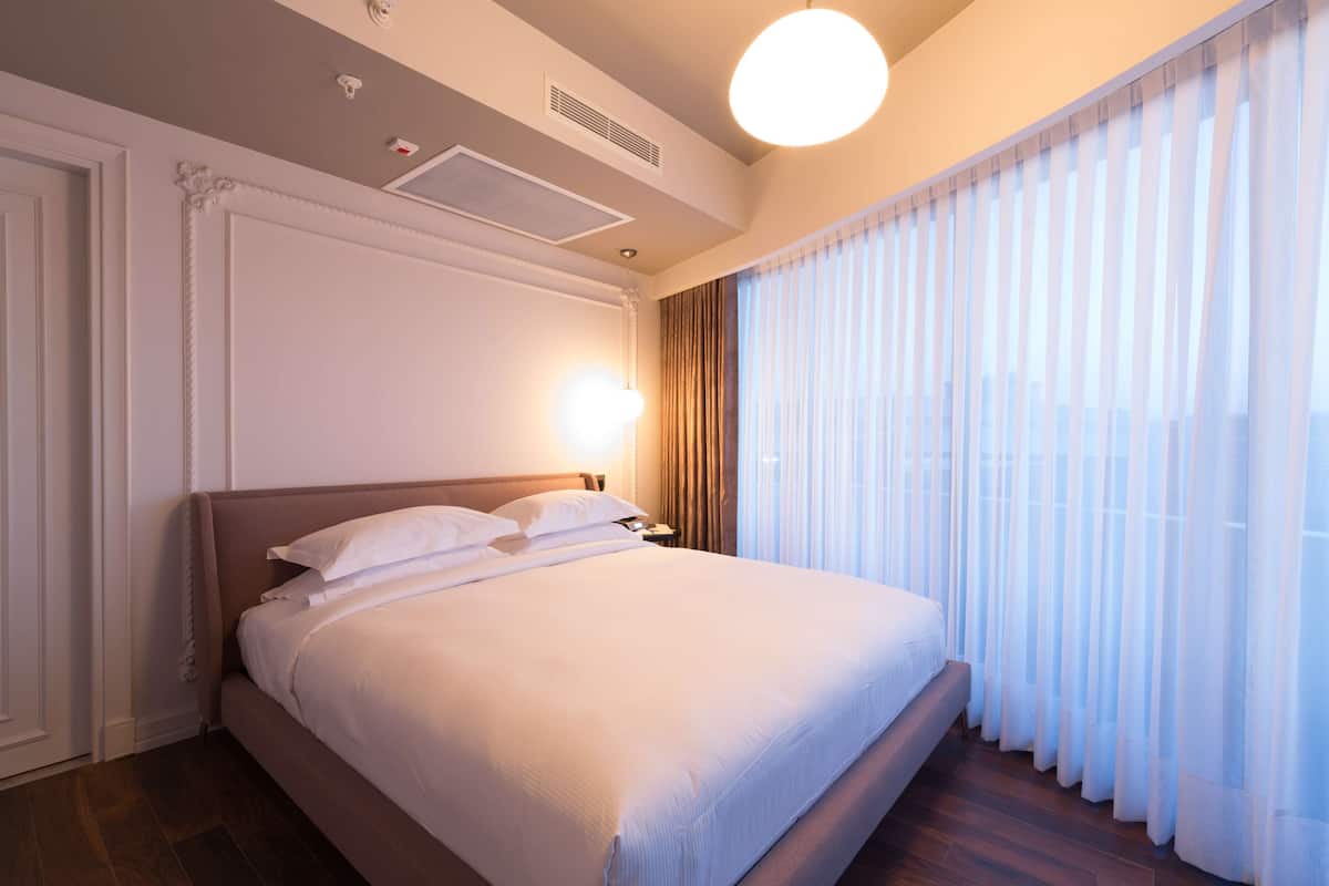 Un lit bien fait, avec du linge de lit blanc, dans une chambre d’hôtel dotée d’une grande fenêtre, d’un plancher en bois et d’un design moderne au plafond.