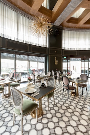 Um restaurante espaçoso com piso em padrão hexagonal, diversas mesas e cadeiras, e amplas janelas com vista para a paisagem urbana.