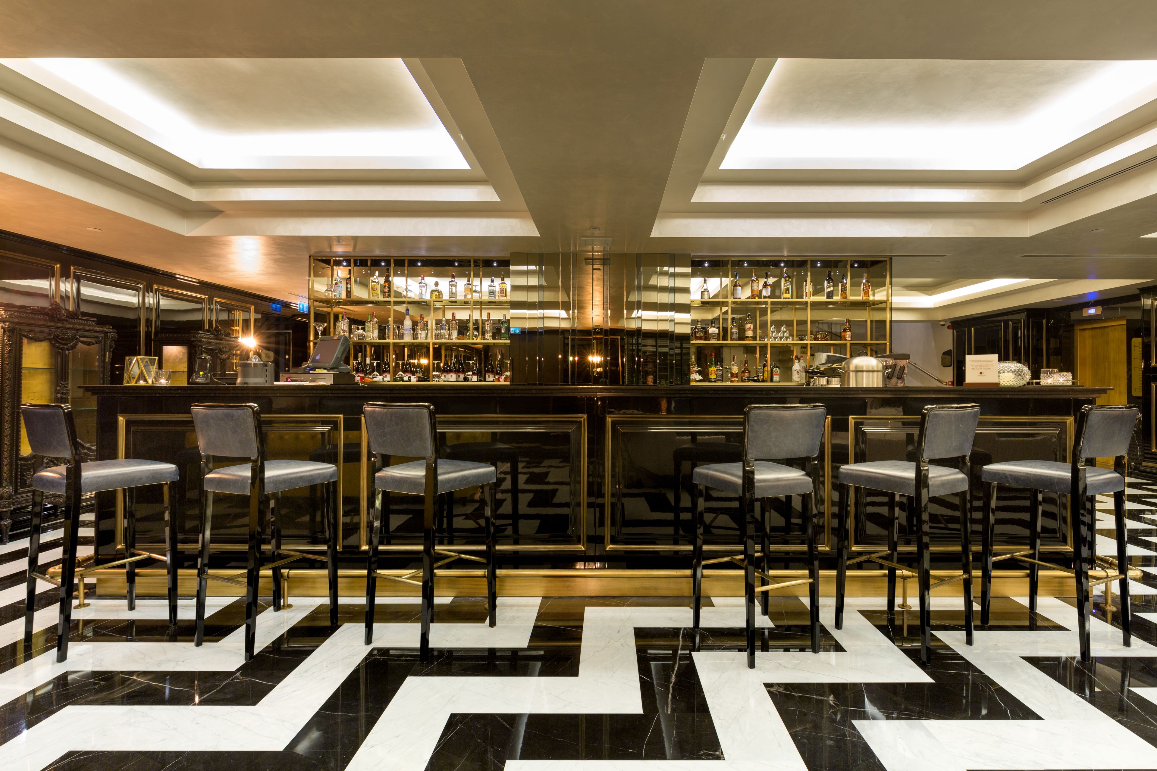 Una zona de bar con suelo de cuadros, taburetes y estanterías llenas de botellas.