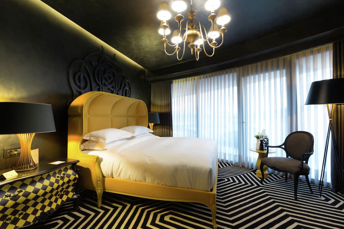 Une chambre d’hôtel avec un grand lit, un lustre, un fauteuil à motifs à carreaux et une table d’appoint avec une lampe.