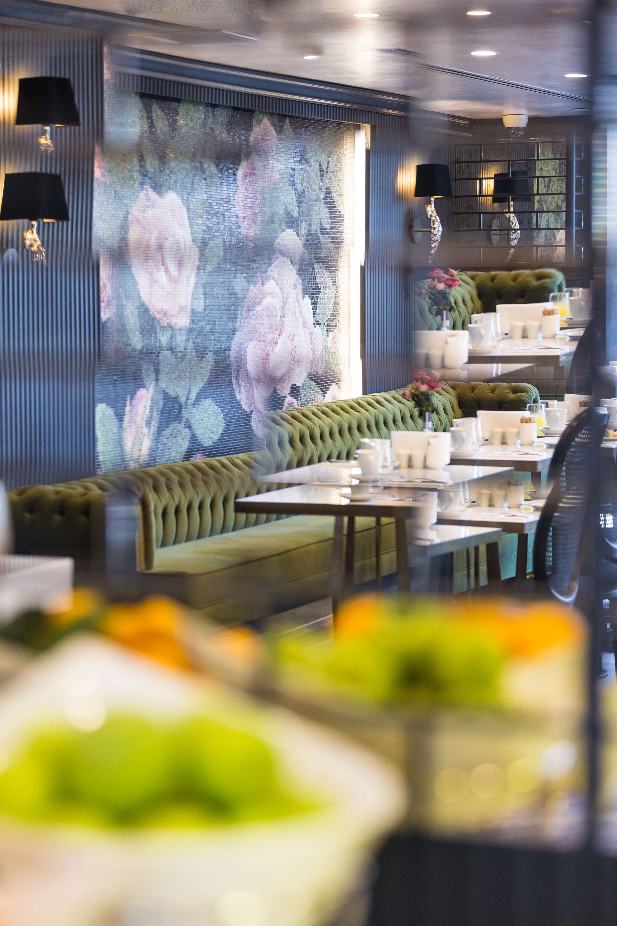Un restaurant avec un banc recouvert d’un tissu vert, des tables avec de la vaisselle blanche et une fresque florale sur le mur.