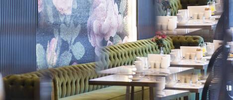 Un restaurant avec un banc recouvert d’un tissu vert, des tables avec de la vaisselle blanche et une fresque florale sur le mur.