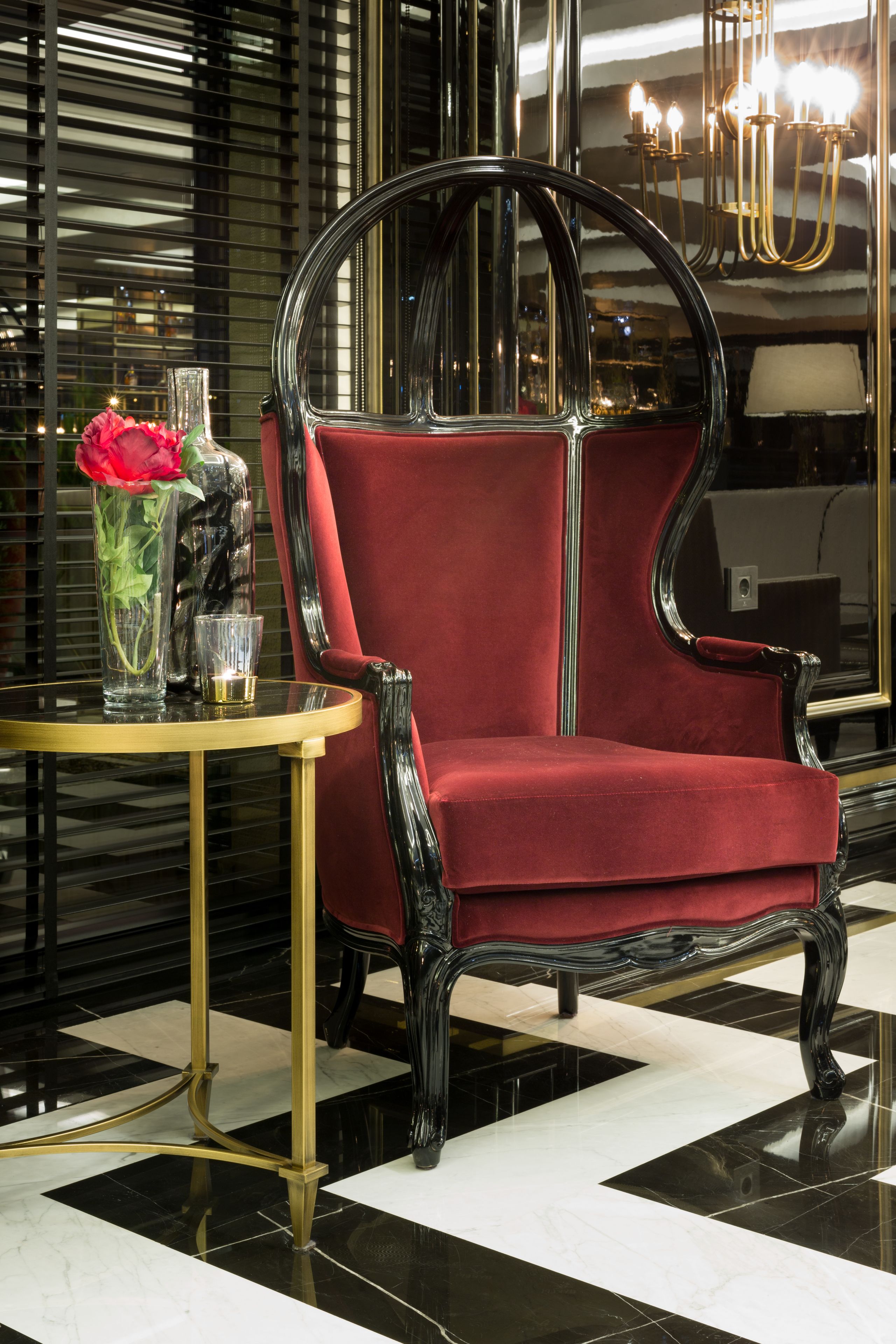 Un sillón tapizado en terciopelo rojo lujoso, con marco dorado, ubicado junto a una mesa de cristal con un florero y un vaso.