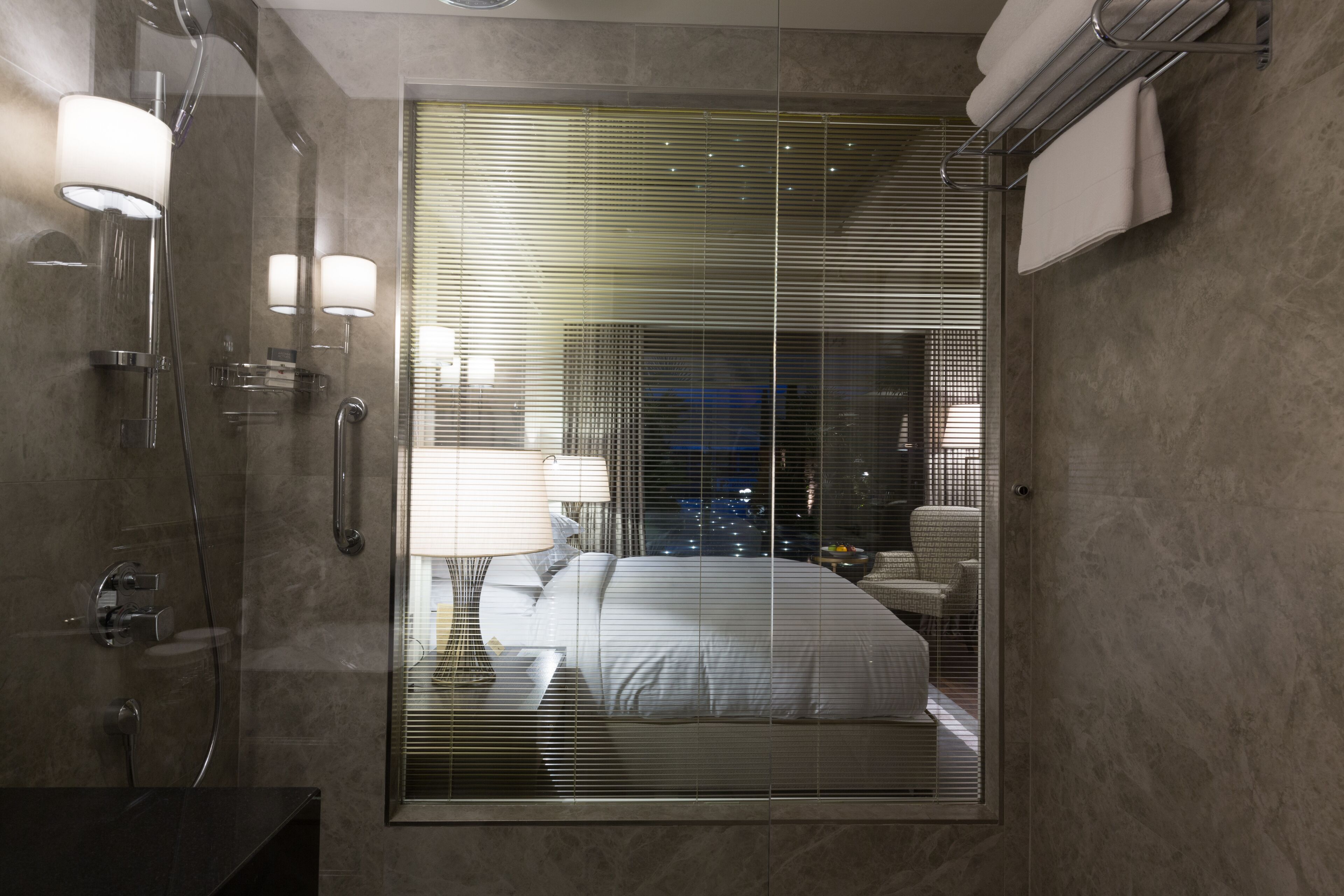 A modern bathroom with a glass-enclosed shower, a visible bed with white bedding, and a towel rack on the wall.