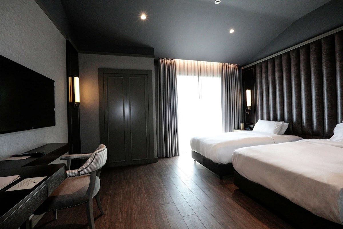 Une chambre d’hôtel équipée d’un téléviseur à écran plat, d’un bureau, de deux lits, d’une chaise et d’une grande fenêtre avec des rideaux.