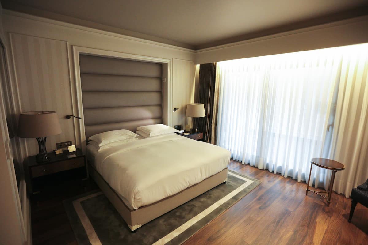 Une chambre d’hôtel avec un grand lit, une table de chevet, une lampe, une petite table et une vue sur l’extérieur à travers des rideaux légers.