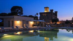 A swimming pool with lounge chairs and umbrellas, a stone building with a tower, and a clear sky at dusk. - Vinilia Wine Resort (Manduria)