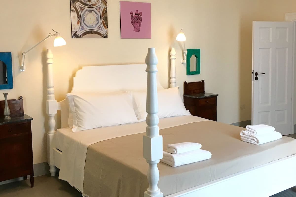 Una cama bien hecha con cabecera blanca, una mesita de noche con lámpara y una pared decorada con dos cuadros.