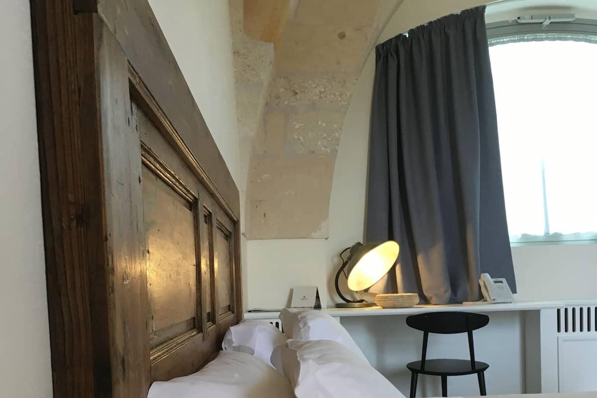 Un dormitorio con cabecera de madera, una lámpara de noche, una cama con ropa blanca, una mesita con lámpara y una ventana con cortinas.